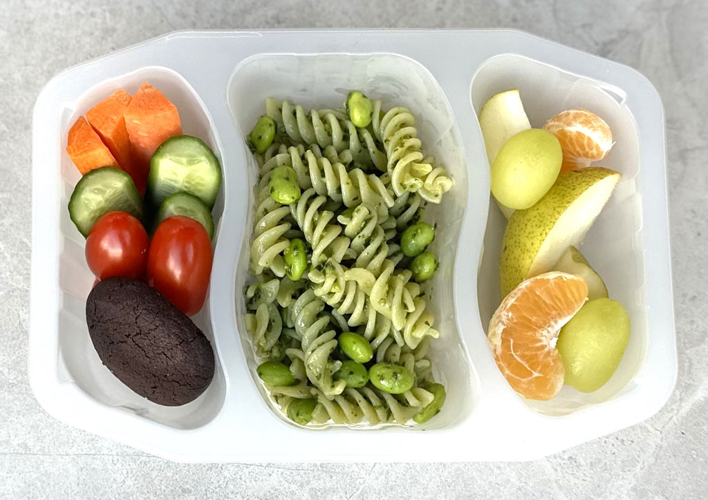 bento style school lunch pack including green pesto spiral pasta with edamame beans, veggie sticks, chocolate cookie and fruit